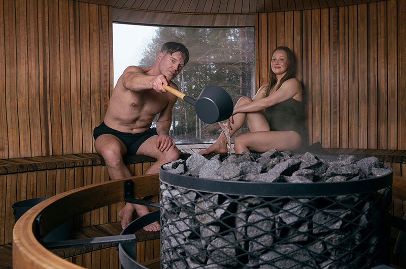 Un homme verse de l'eau sur les pierres chauffées du sauna avec une louche, une femme est assise à proximité ; un sauna rond en bois fumant avec une fenêtre.