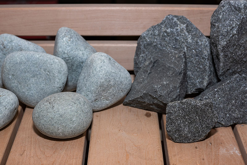 Comparaison : pierres de rivière arrondies de couleur claire et pierres de sauna foncées brisées placées sur un banc en bois.